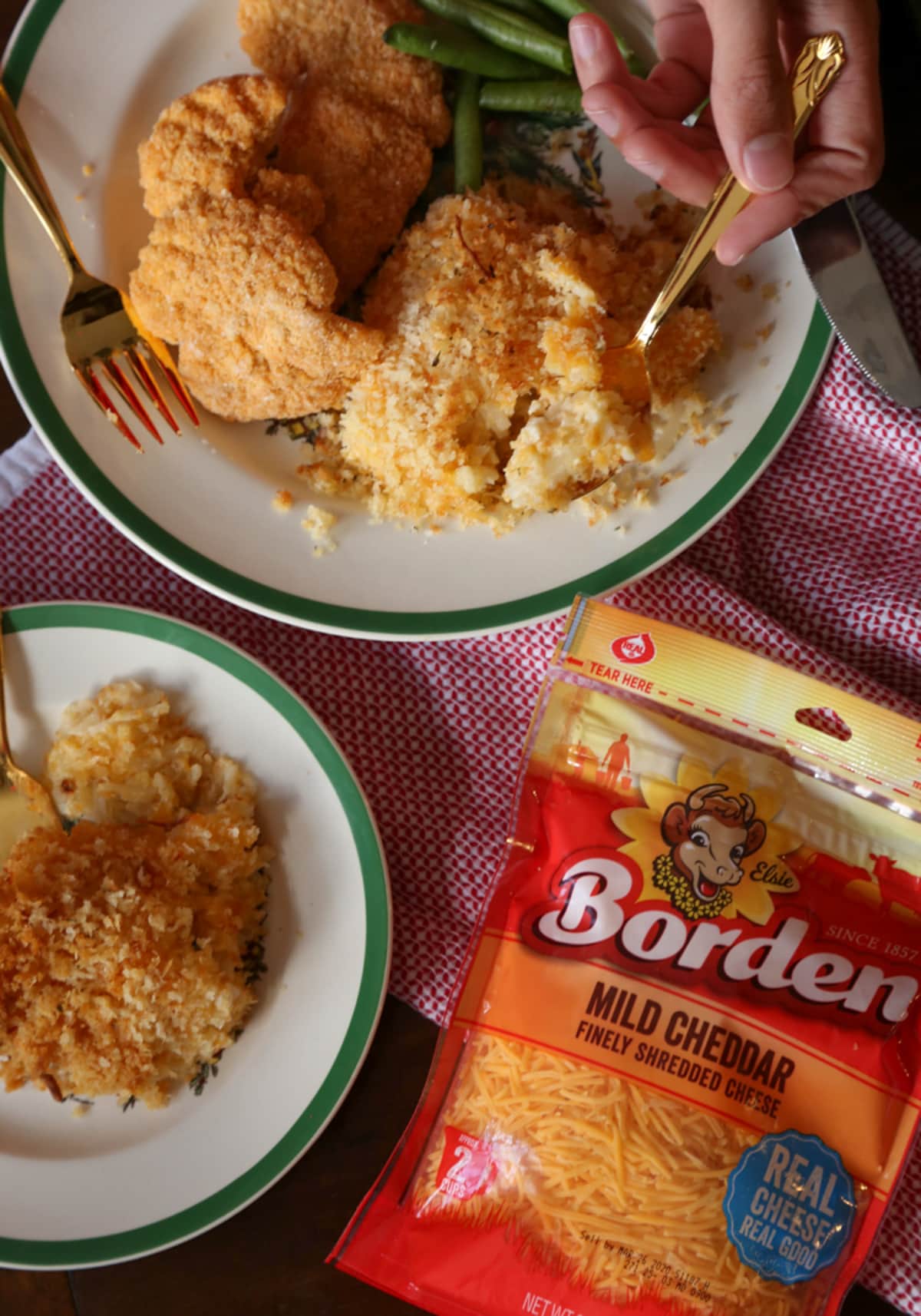 Overhead view of a plate with green beens, chicken, and casserole, with a hand holding a spoon, next to a pack of cheese and a bowl of casserole.