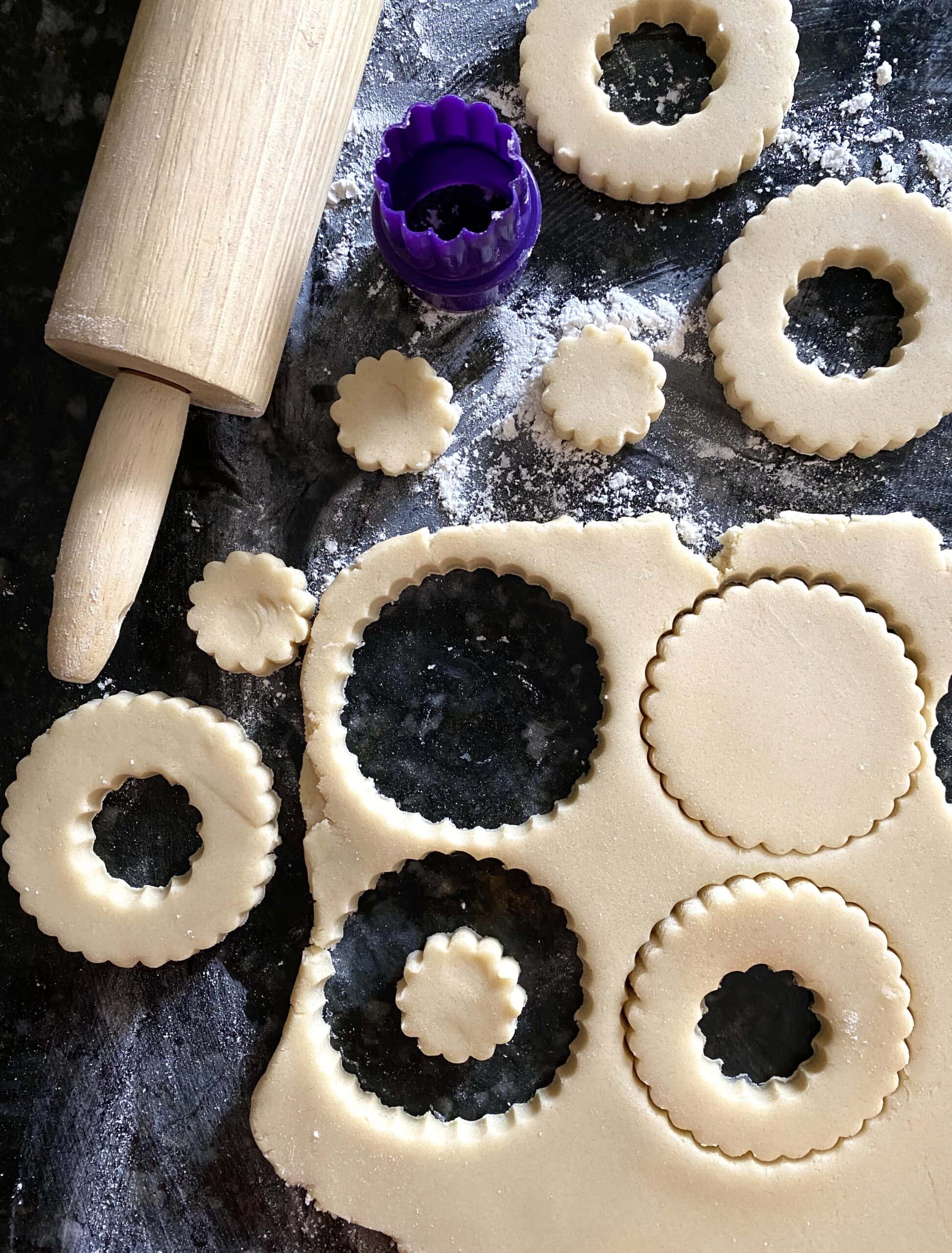 Overhead view of Christmas wreath cookies partially cut out from rolled out sugar cookie dough, on a floured countertop next to a rolling pin.