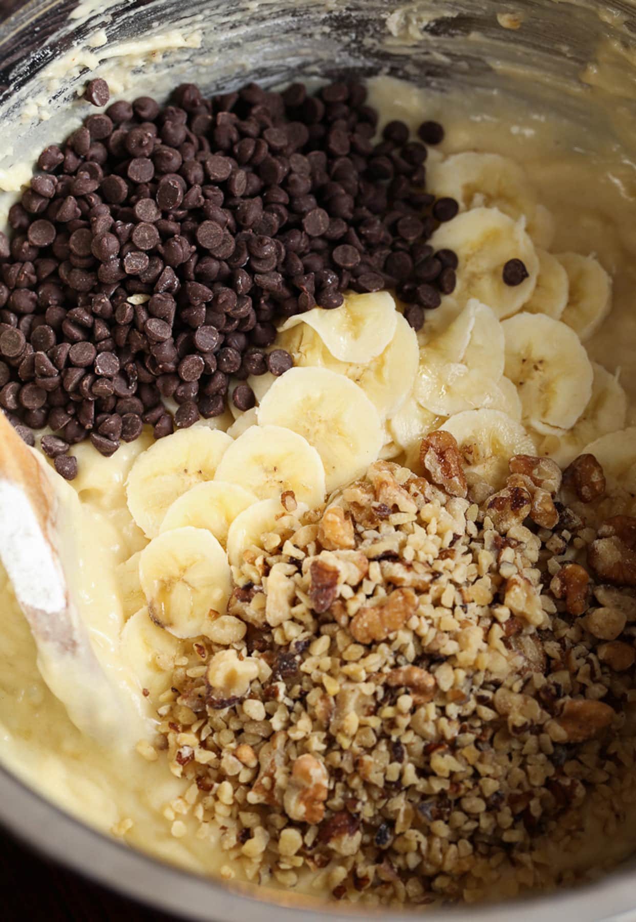 Banana bread batter in a mixing bowl with chocolate chips, chopped walnuts, and sliced bananas to be folded in.