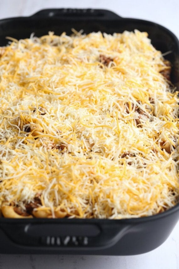 Chili mac ingredients topped with shredded cheese in a large black casserole dish.