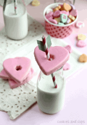 A heart-shaped Valentine's cookie on top of a glass of milk with a straw through the center.