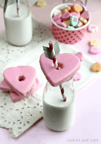 A heart-shaped Valentine's cookie on top of a glass of milk with a straw through the center.