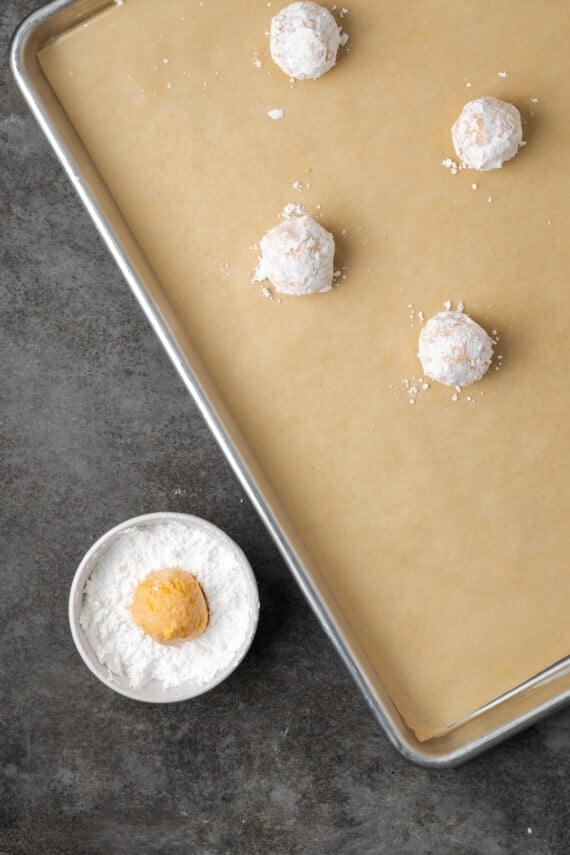 An orange cookie dough ball in a small bowl of powdered sugar, next to more dough balls coated in sugar arranged on a lined baking sheet.