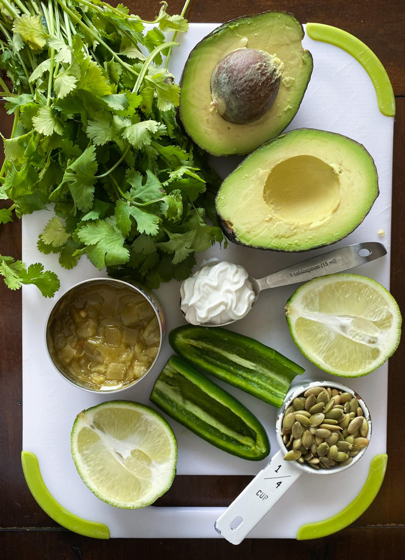 A cutting board with cilantro, avocado, green chilis, limes, jalapenos, sour cream, and pumpkin seeds to make avocado salsa