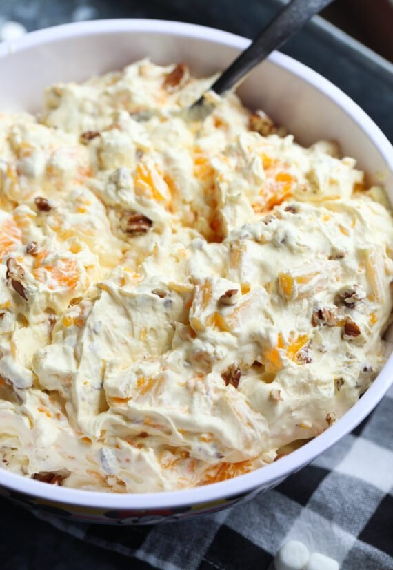 A close up image of bowl of orange cream fruit salad topped with chopped pecans.