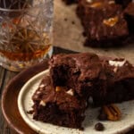 Bourbon pecan brownies stacked on a plate, with a bite missing from the top brownie and more brownies next to a glass of bourbon in the background.