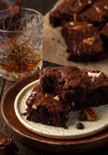 Bourbon pecan brownies stacked on a plate, with a bite missing from the top brownie and more brownies next to a glass of bourbon in the background.