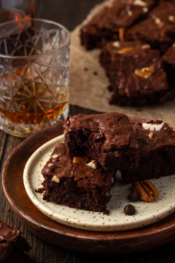 Bourbon pecan brownies stacked on a plate, with a bite missing from the top brownie and more brownies next to a glass of bourbon in the background.