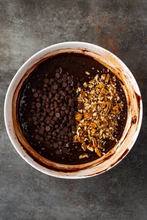 Chopped pecans and chocolate chips added to brownie batter in a white bowl.