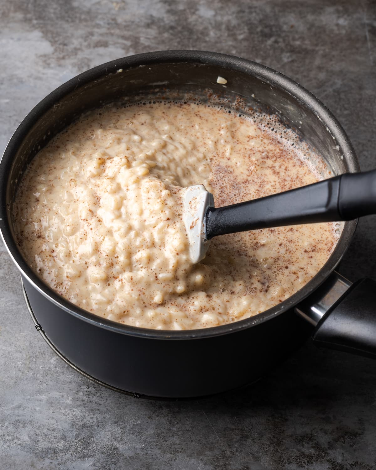 A black saucepan filled with creamy rice pudding with a spatula.