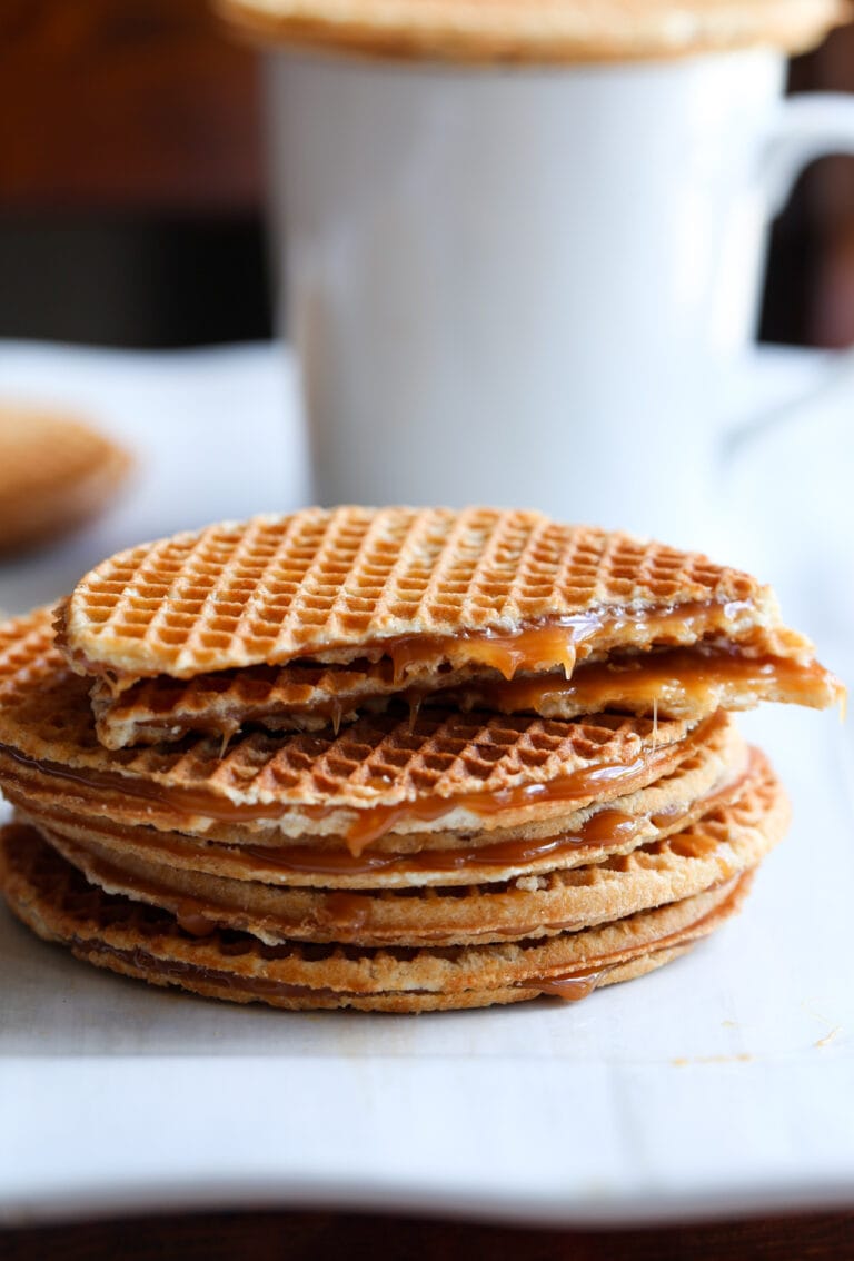 Stroopwafels stacked on a plate next to a cup of coffee, with the top cookie broken in half.