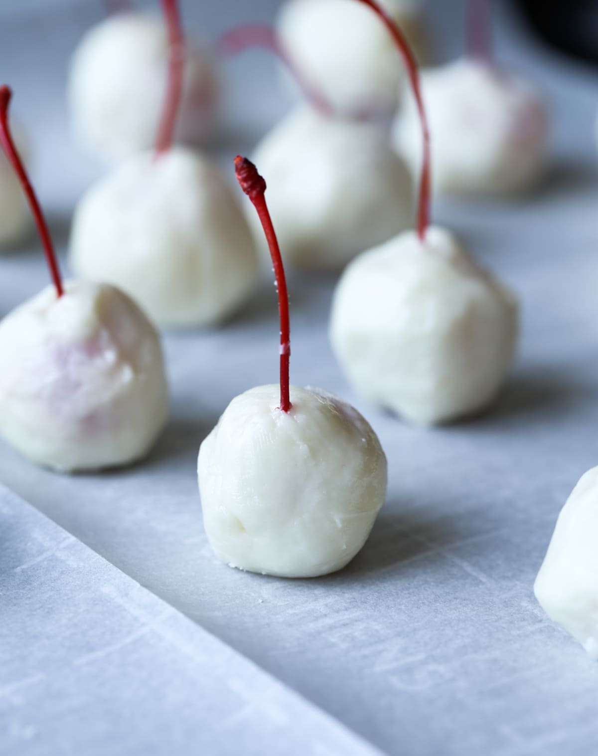 Cherries with a stem coated in white fondant on a parchment lined baking sheet