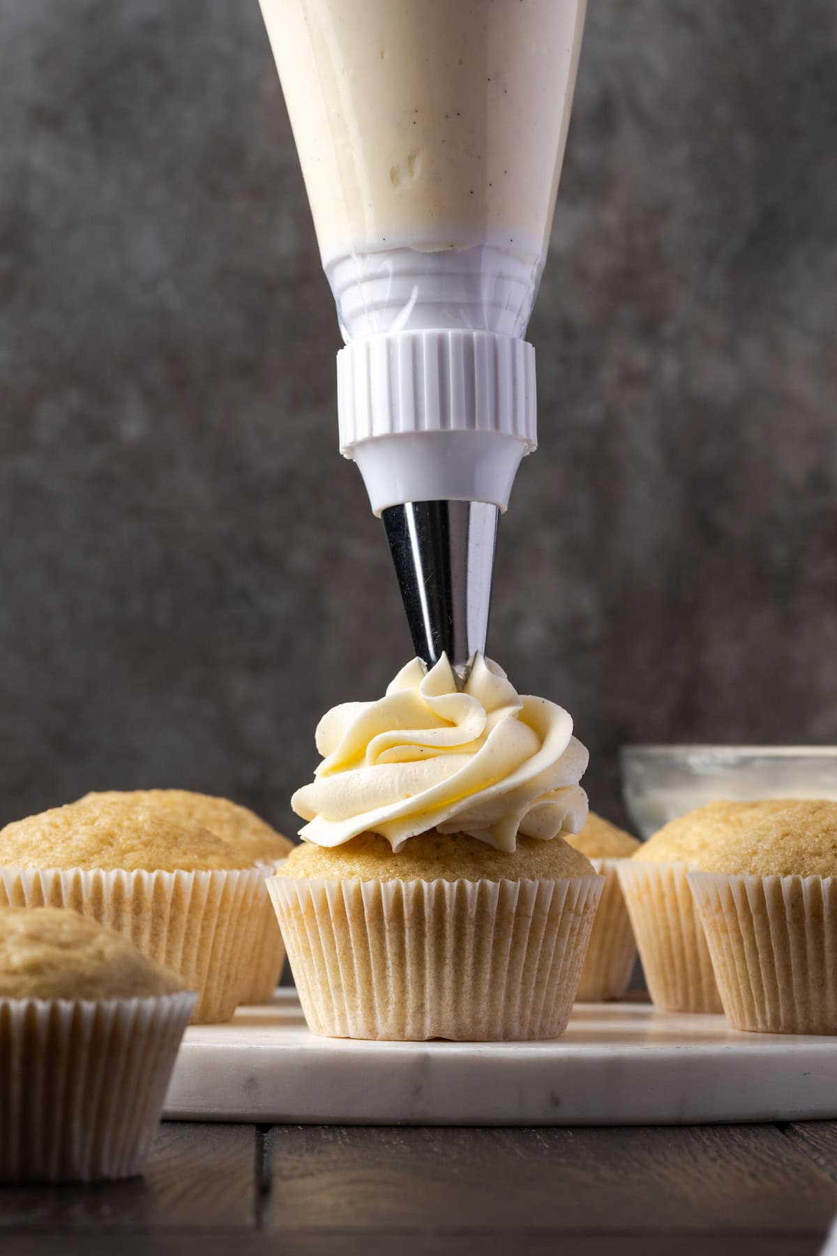 A piping bag fitted with a piping tip piping a swirl of buttercream onto a cupcake, with more unfrosted cupcakes int he background.