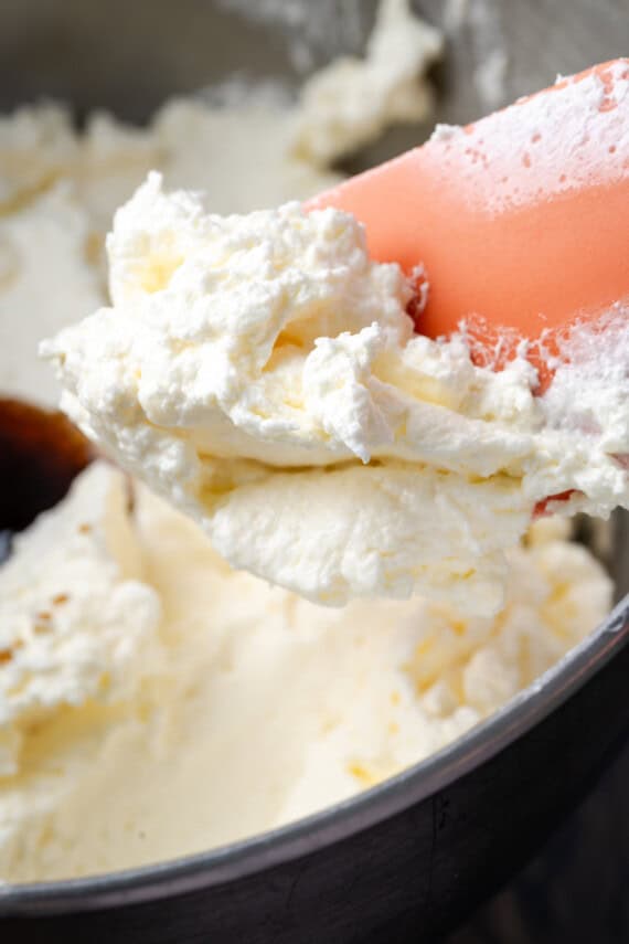 An orange rubber spatula holding a scoop of Swiss meringue buttercream when the butter has just been added, over a bowl of frosting.