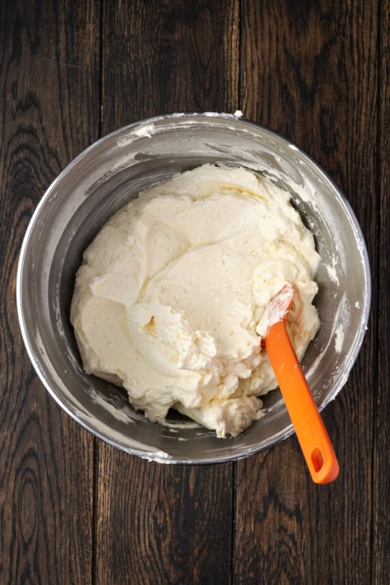 An orange rubber spatula resting in a bowl of buttercream frosting.