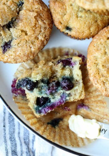 Overhead view of one half of a banana blueberry muffin resting in a muffin liner, next to more muffins on a plate.