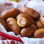 A mini corn dog with a bite taken out of it, in a basket of more corn dogs.