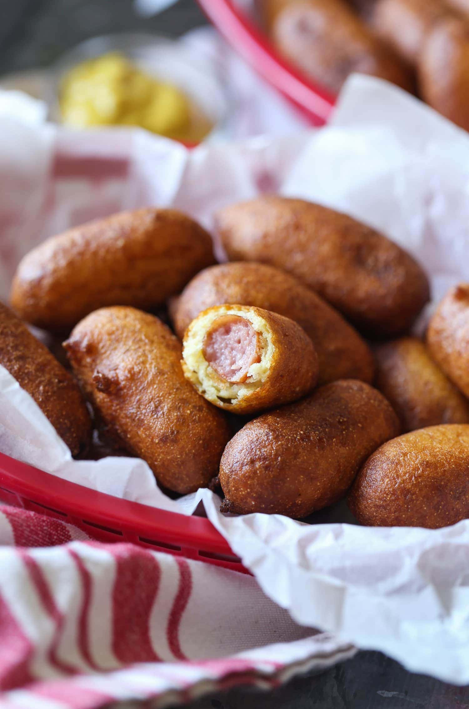 A mini corn dog with a bite taken out of it, in a basket of more corn dogs.