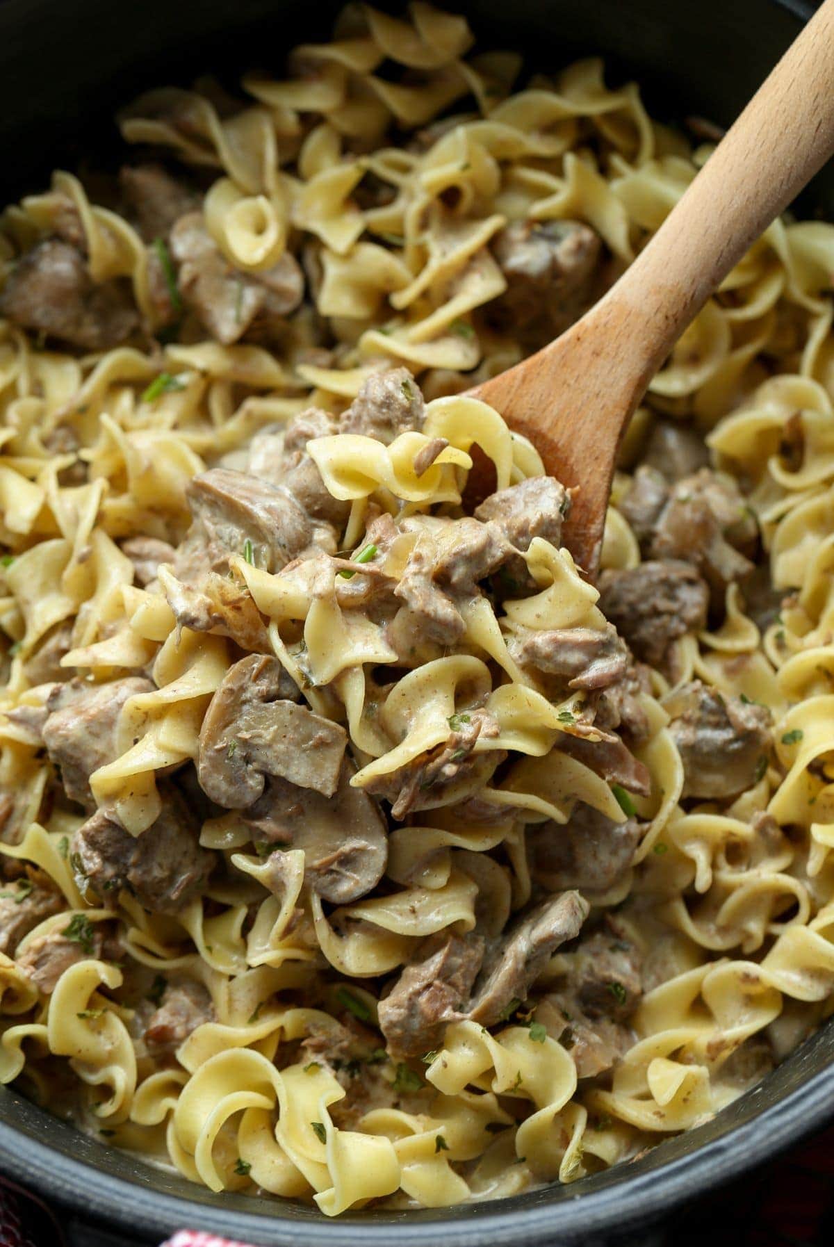 A portion of stroganoff with egg noodles being scooped out of a Crockpot with a wooden spoon.