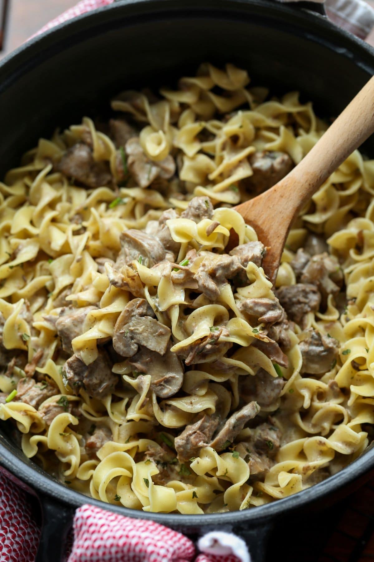 A Crockpot filled with beef stroganoff and egg noodles. A wooden spoon is lifting out a spoonful.