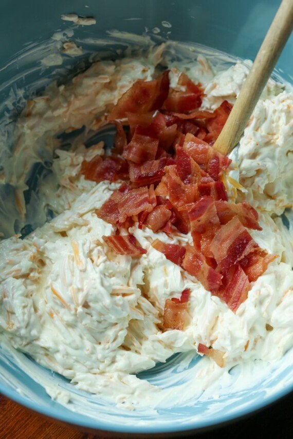 A mixing bowl with a mixture of cream cheese, sour cream, cheddar, and chopped bacon being stirred together in it.