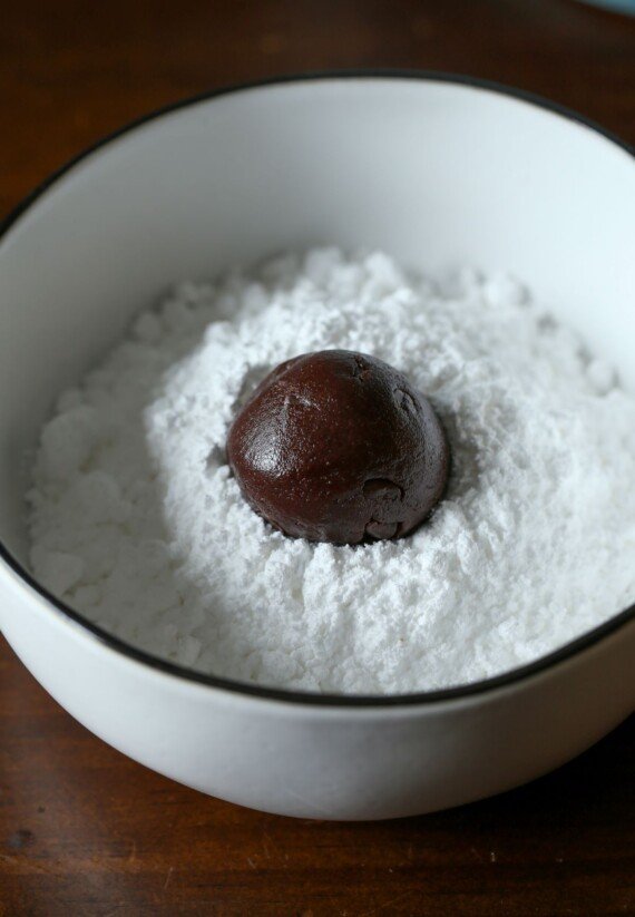 A white enamel bowl of powdered sugar with one ball of shiny, chocolate cookie dough in the sugar.