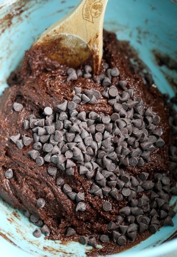 A bowl of chocolate cookie dough with chocolate chips piled on top. A wooden spoon is partially submerged in the dough.
