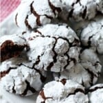 Freshly baked chocolate crinkle cookies on a white tea towel and gingham tablecloth. One of the cookies has been broken in half to reveal the soft interior.