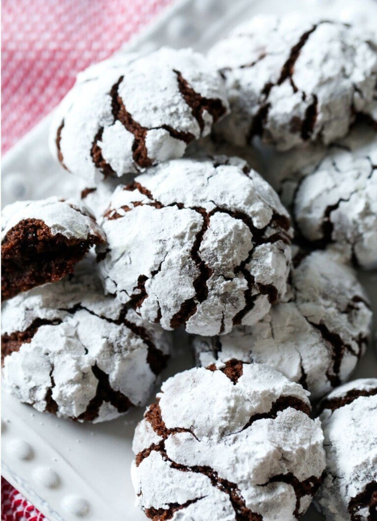 Freshly baked chocolate crinkle cookies on a white tea towel and gingham tablecloth. One of the cookies has been broken in half to reveal the soft interior.