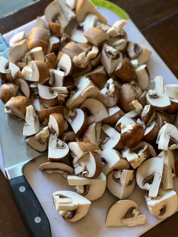 A cutting board with a knife and a pile of raw, quartered baby bella mushrooms.