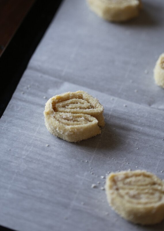 Slices of folded and cut palmier cookie dough are placed cut-side-up on a baking sheet.