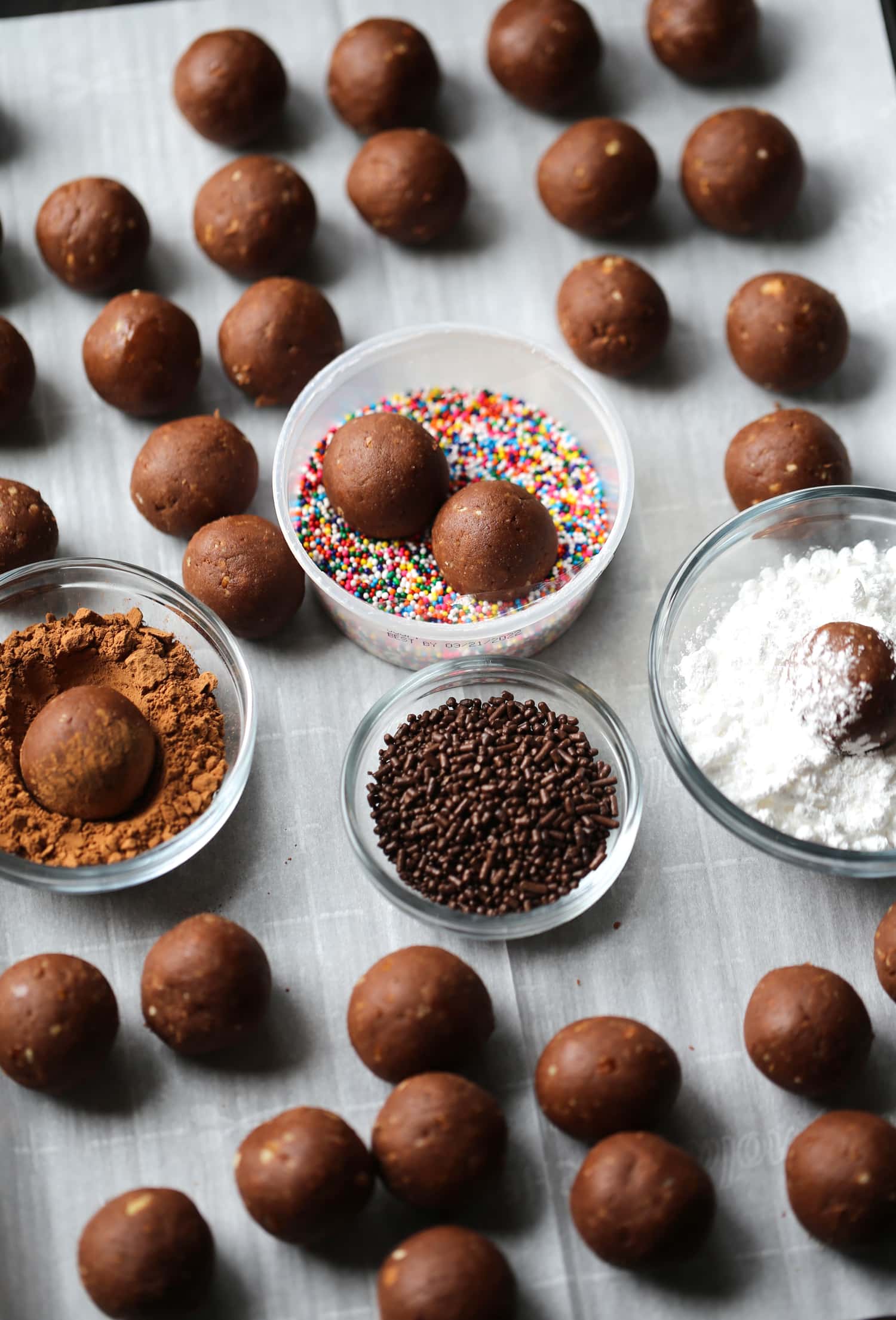 An assortment of holiday rum balls being rolled in bowls of cocoa powder, sprinkles, and powdered sugar.