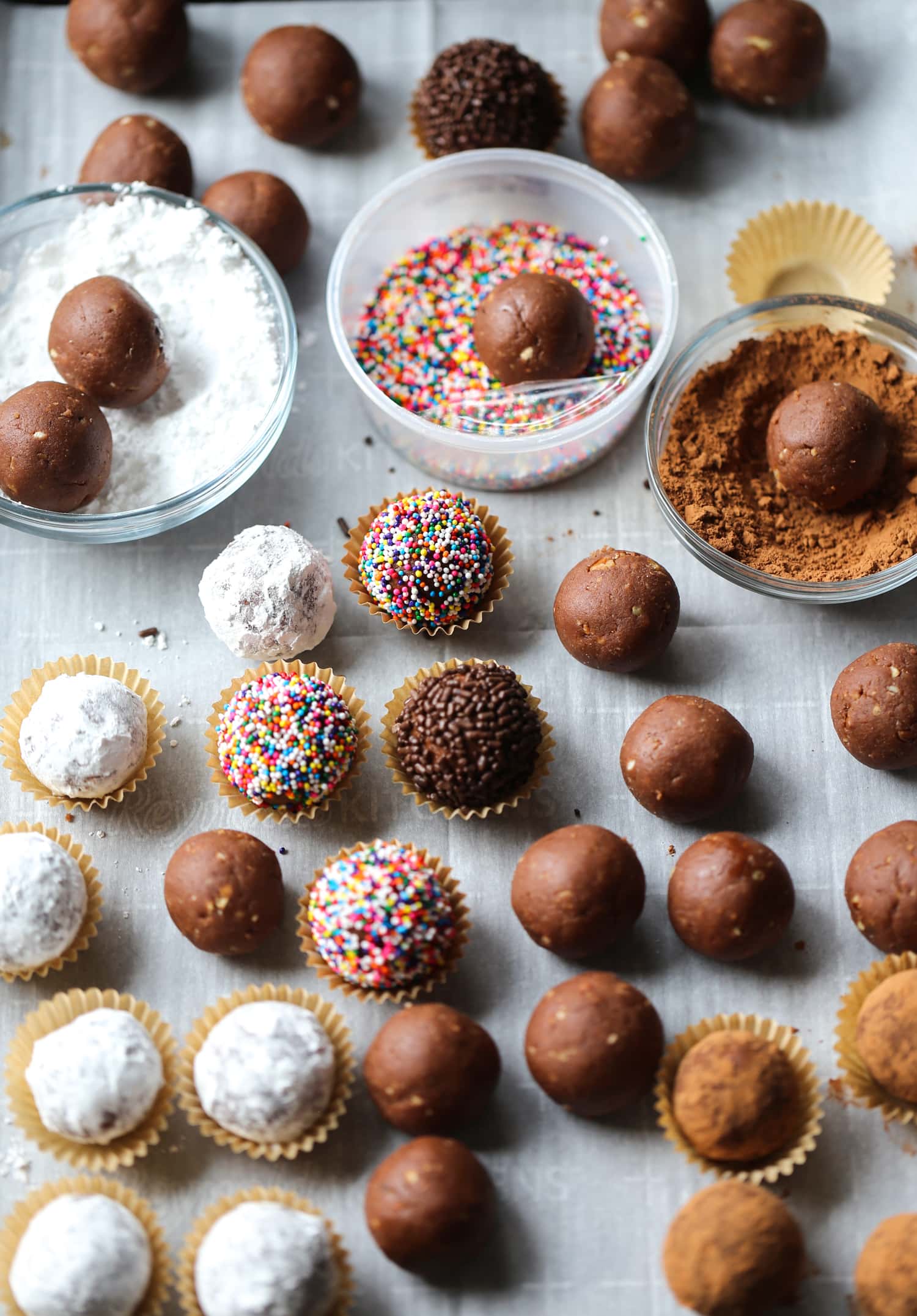 An assortment of holiday rum balls rolled in sprinkles, cocoa powder and powdered sugar, with bowls of decorations in the background.