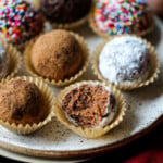Decorated holiday rum balls in candy cups, arranged on a plate, one with a bite missing.