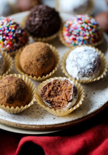 Decorated holiday rum balls in candy cups, arranged on a plate, one with a bite missing.