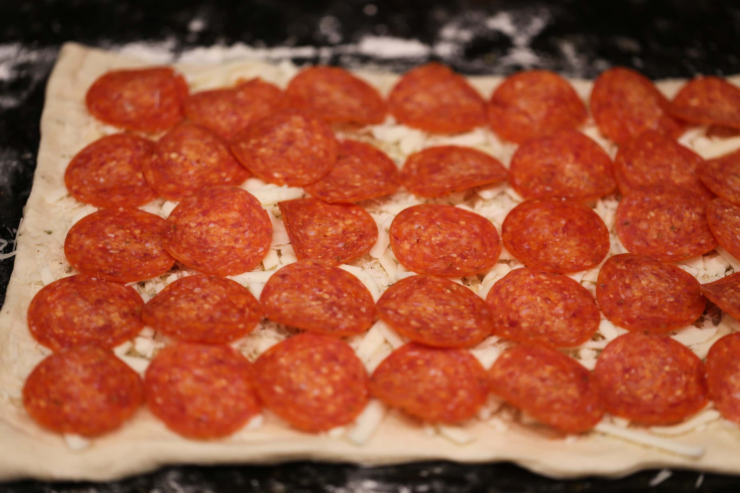 Rolled out pizza dough is topped with cheese and pepperoni.