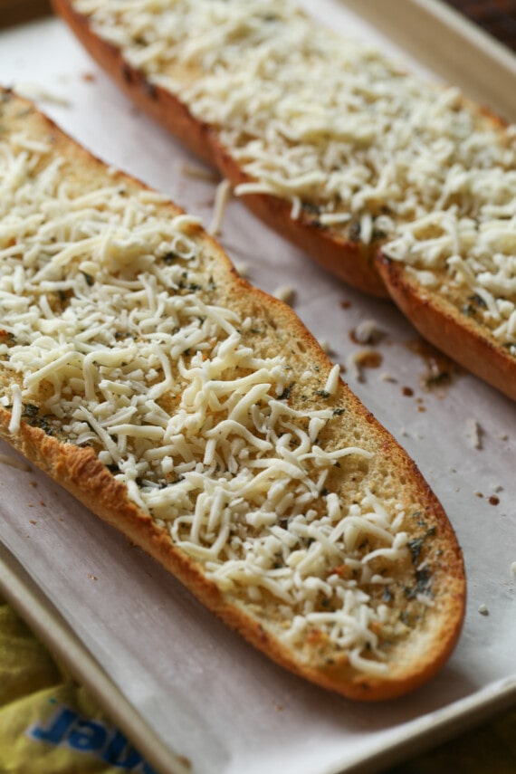 Two halves of a French bread loaf on a baking sheet, topped with a thin layer of shredded cheese.