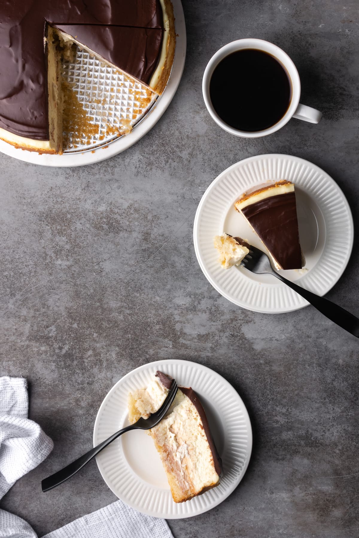A table spread with Boston Cream Pie cheesecake slices on plates, next to a cup of coffee and the remaining cake.