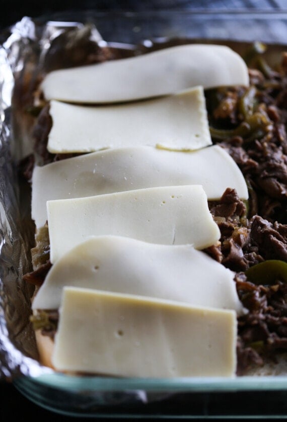 Cheese slices are layered over steak on top of buns in a baking dish.
