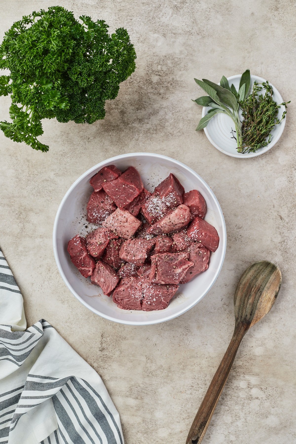 Beef chunks are seasoned in a bowl next to a wooden spoon and glasses of fresh herbs.