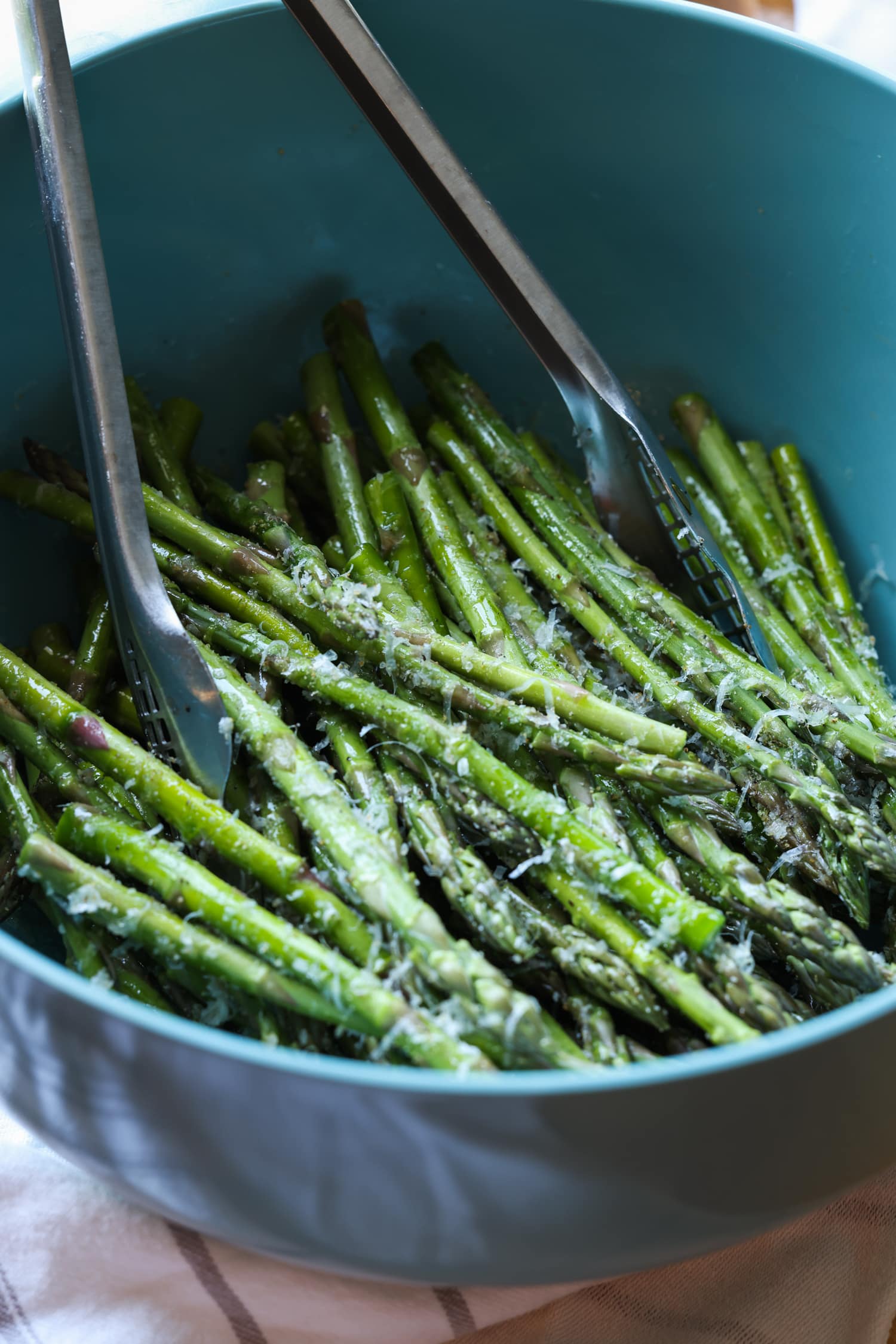 Asparagus spears are tossed with seasonings in a large blue bowl with metal tongs.
