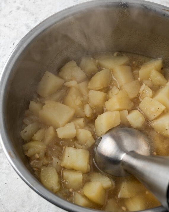 An immersion blender is used to puree potatoes in a pot.
