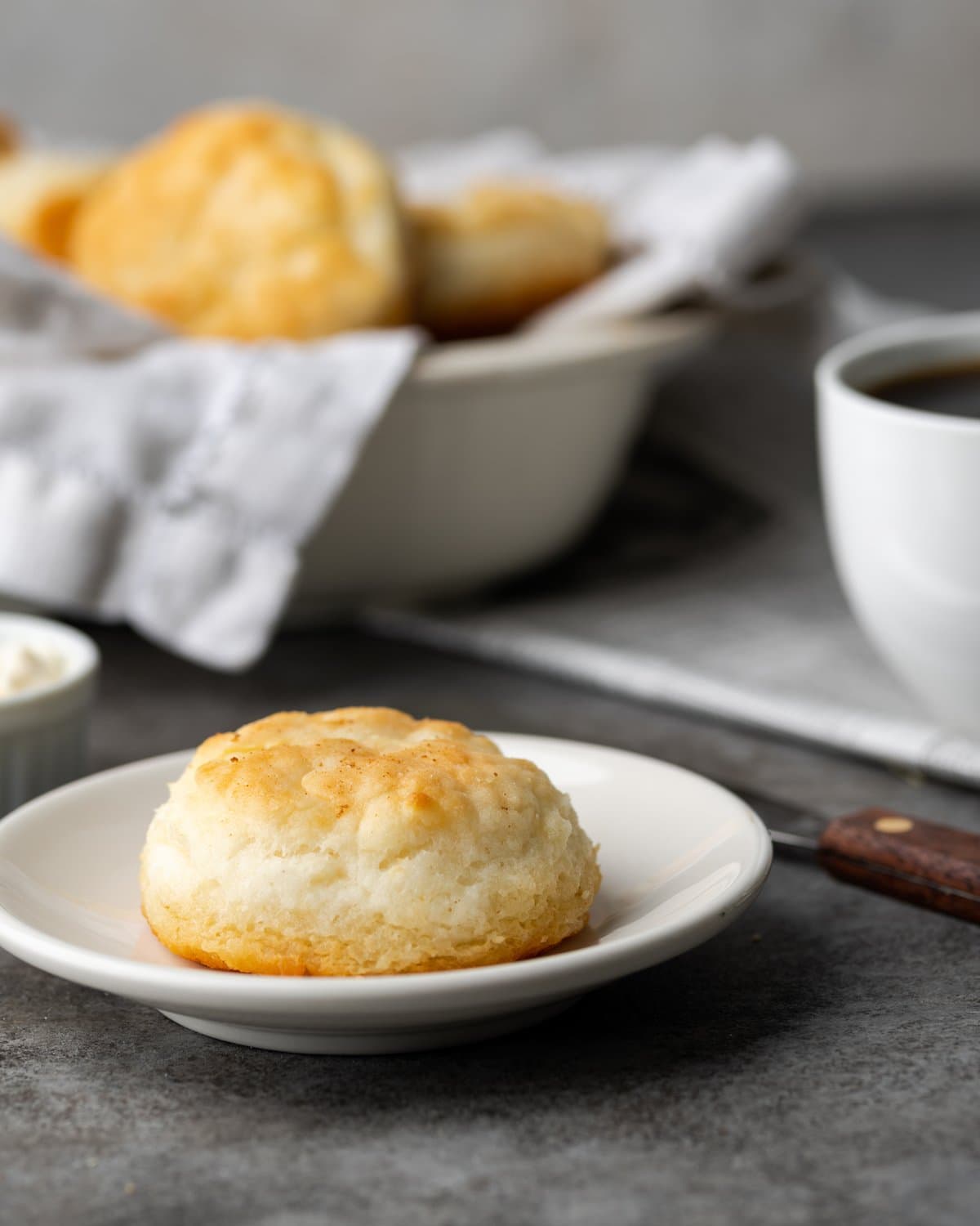 A 7UP biscuit on a white plate, next to a cup of coffee with a bowl of 7UP biscuits in the background.