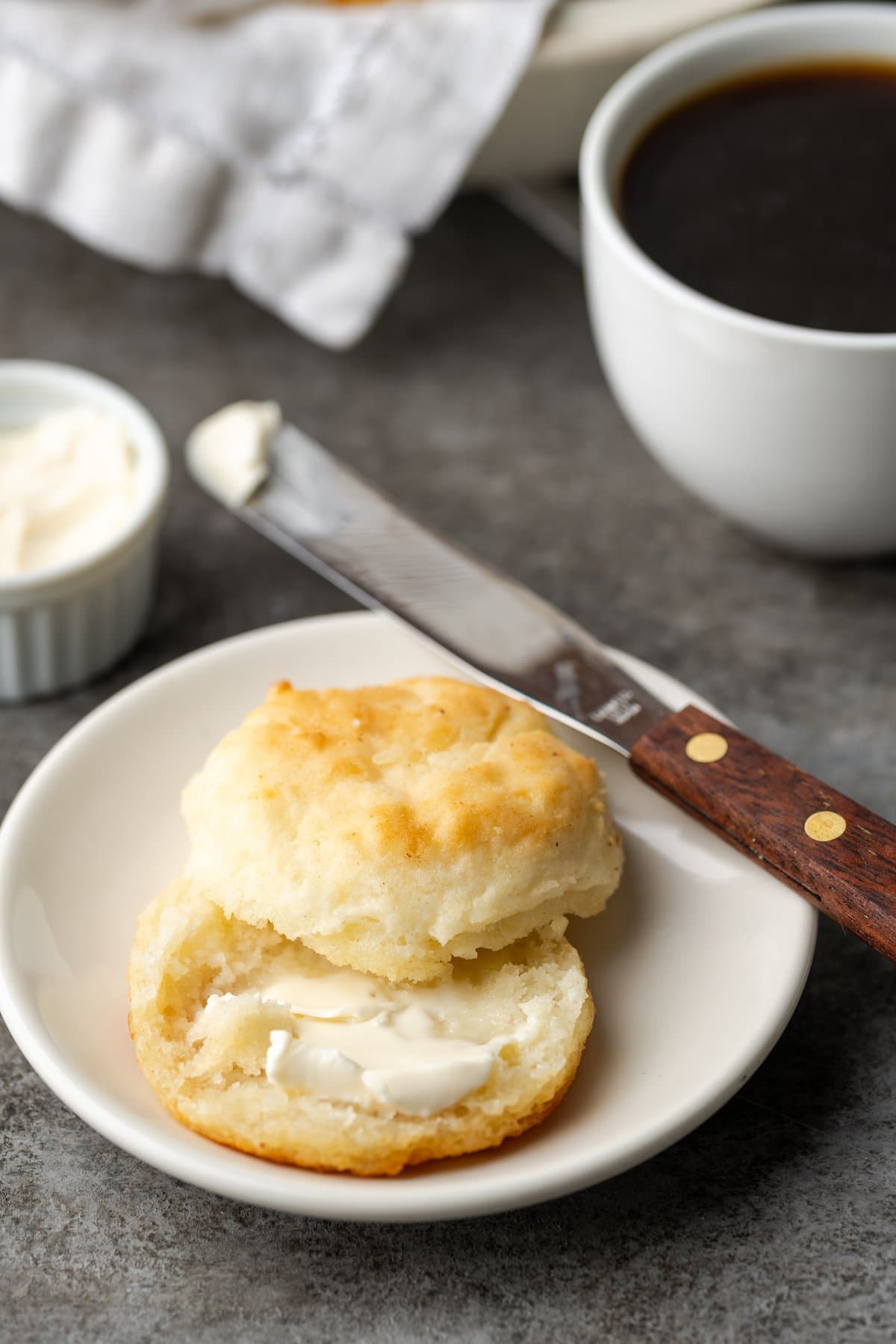 A 7UP biscuit cut in half on a plate next to a butter knife, with one half spread with butter.