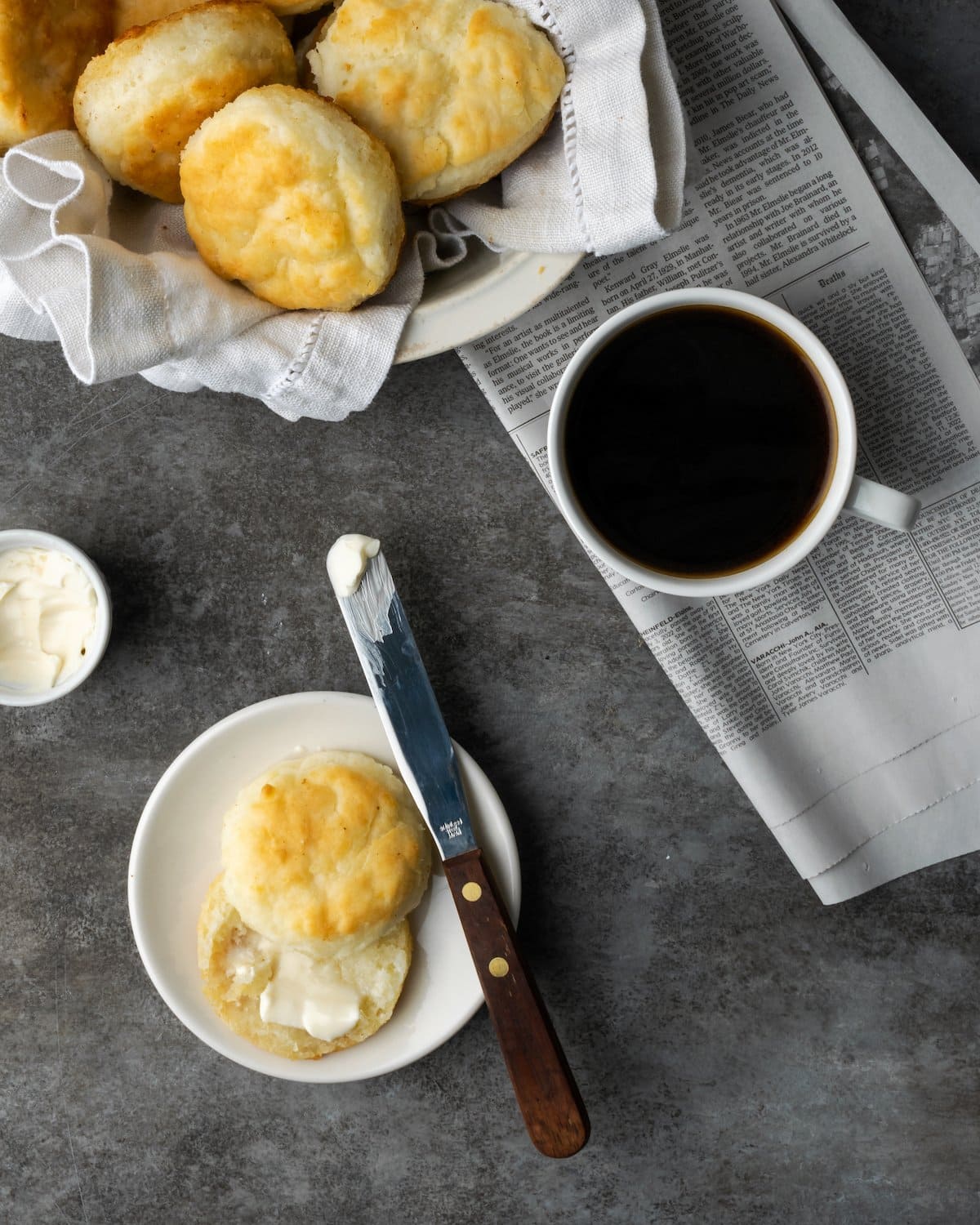 Top view of a breakfast spread with two buttered halves of a 7UP biscuit on a plate next to a cup of coffee and a bowl of biscuits.