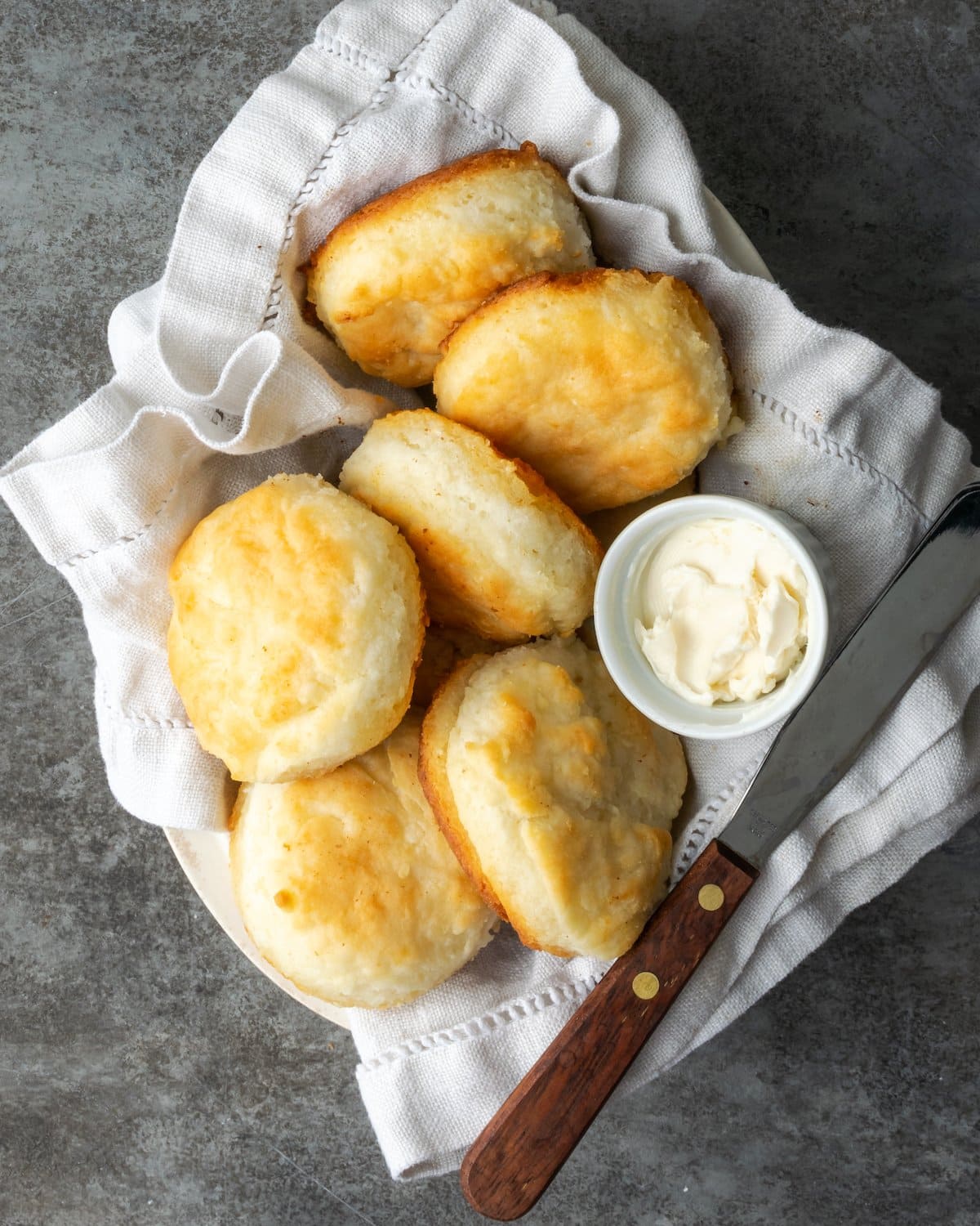 Top view of baked 7UP biscuits in a cloth-lined dish, with a small bowl of butter and a butter knife.