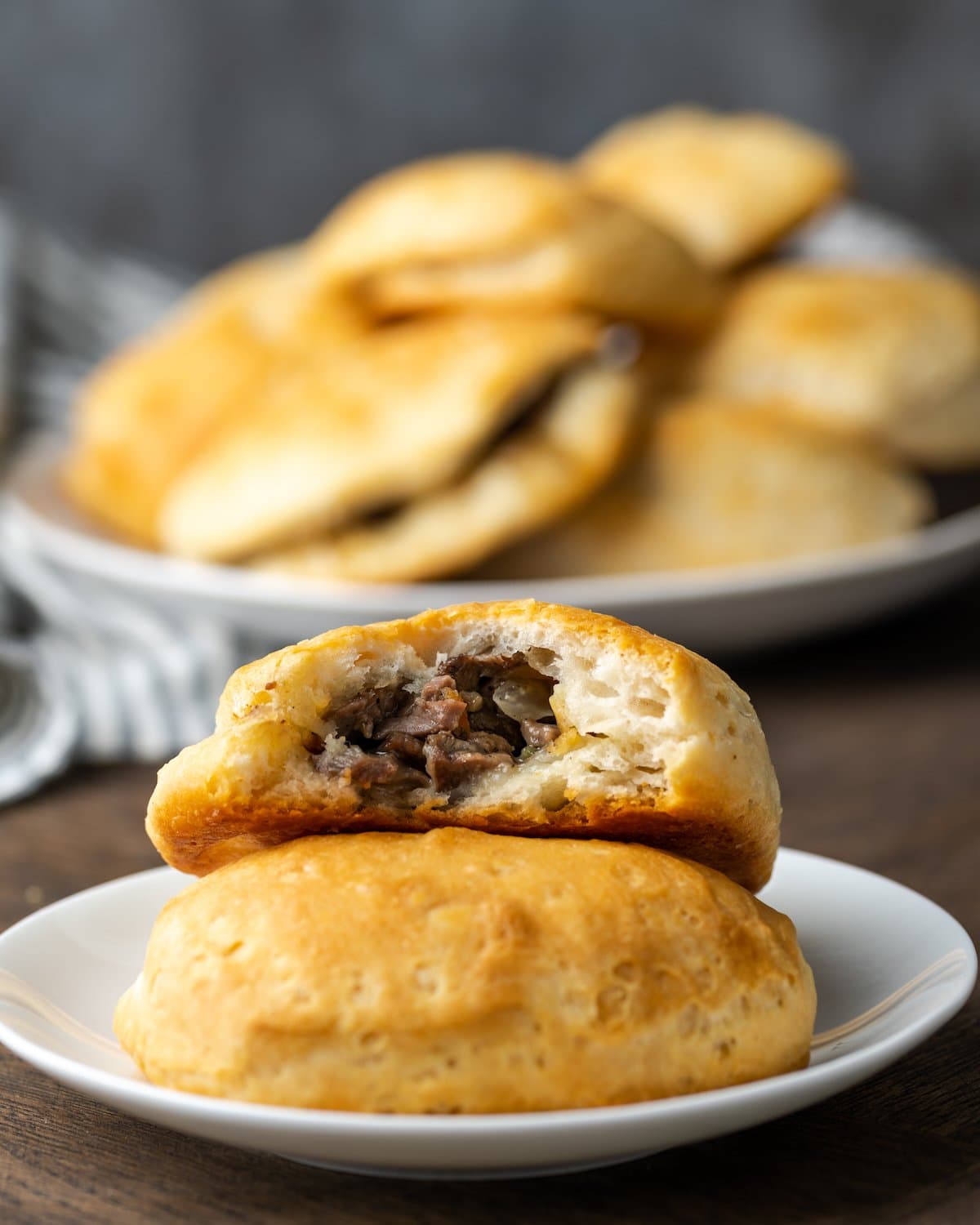 Two Philly cheesesteak stuffed biscuits on a plate, one with a bite missing, with more biscuits in the background.