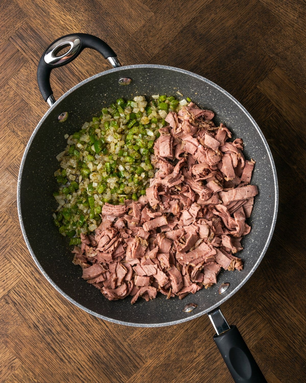 Sliced roast beef added to a skillet with sautéed diced onions and peppers.