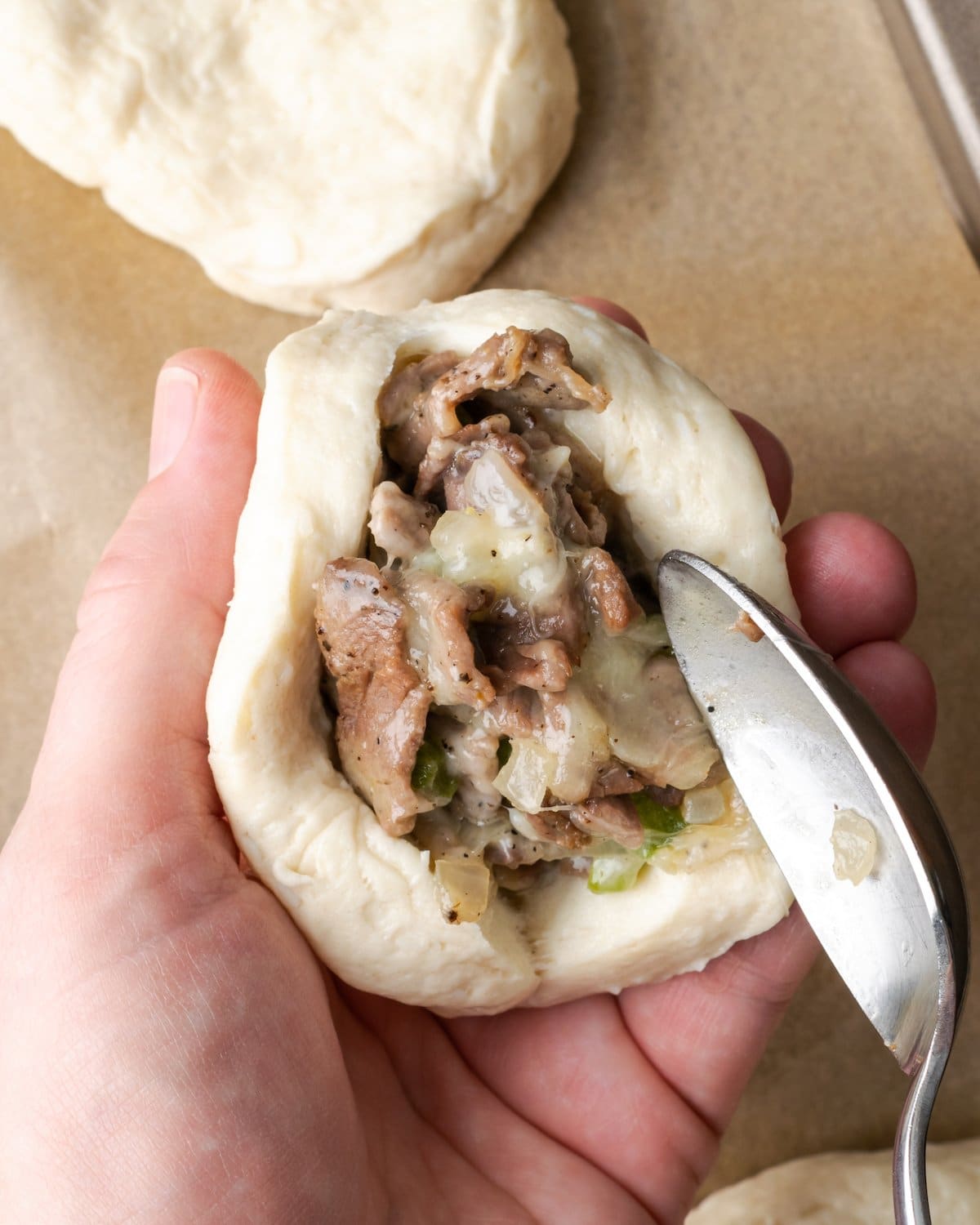 A hand holding biscuit dough while the other hand uses a spoon to fill the dough with Philly cheesesteak.