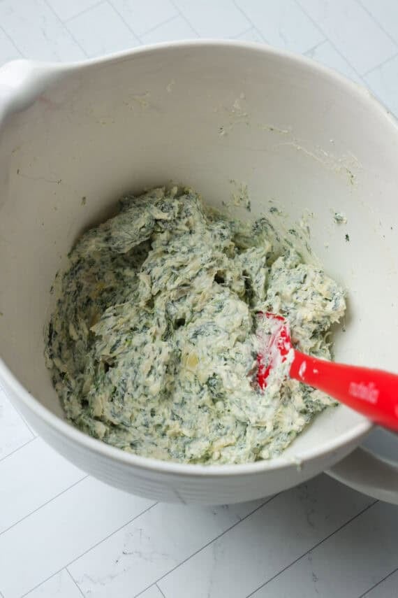 A white mixing bowl with the mixed ingredients for spinach artichoke dip.